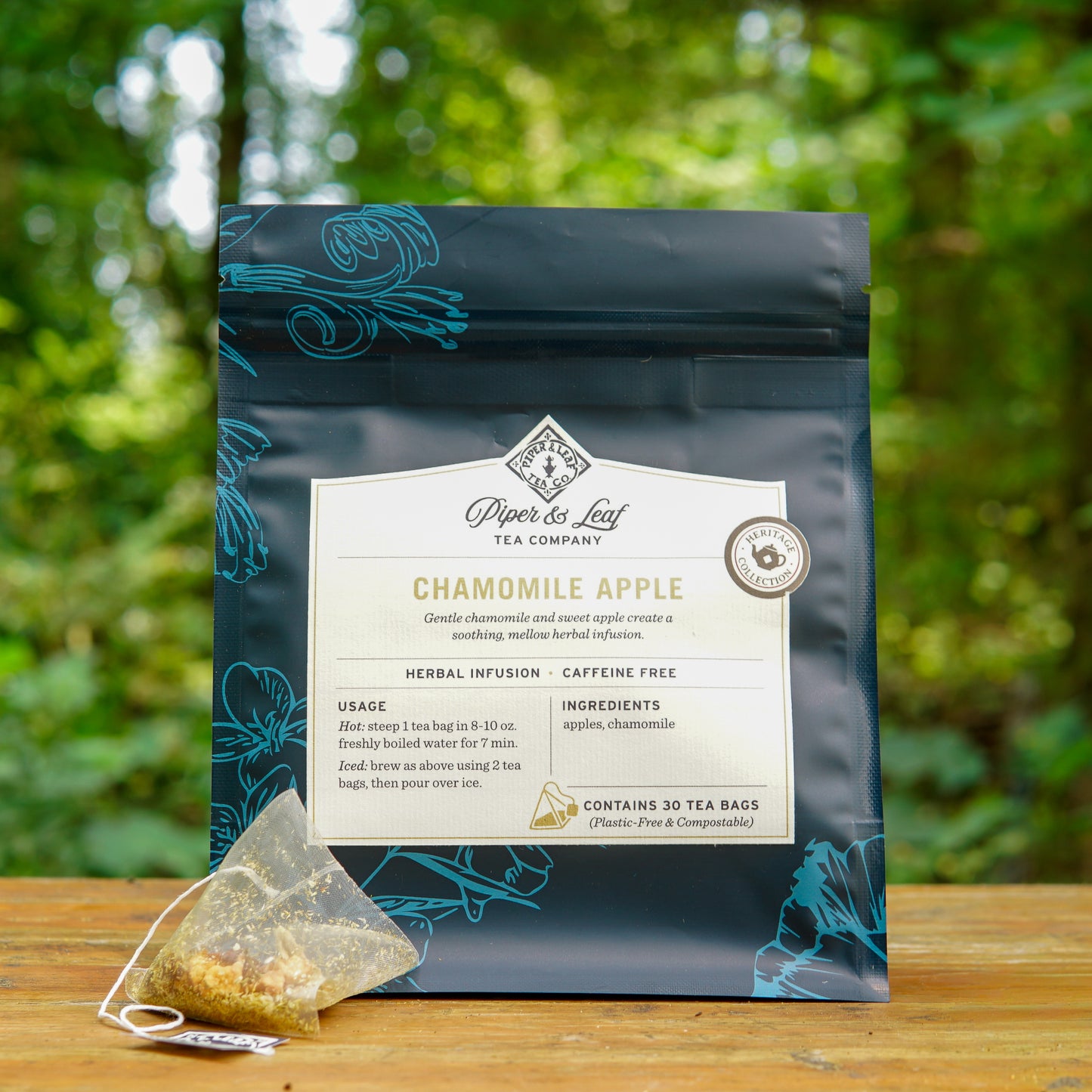 A bag of Piper and Leaf Tea Co. Chamomile Apple 30ct Tea Bags with a tea bag in front, pictured outdoors against a softly blurred green forest backdrop. Savor this caffeine-free herbal blend, crafted with plastic-free tea bags for an eco-friendly and calming experience.