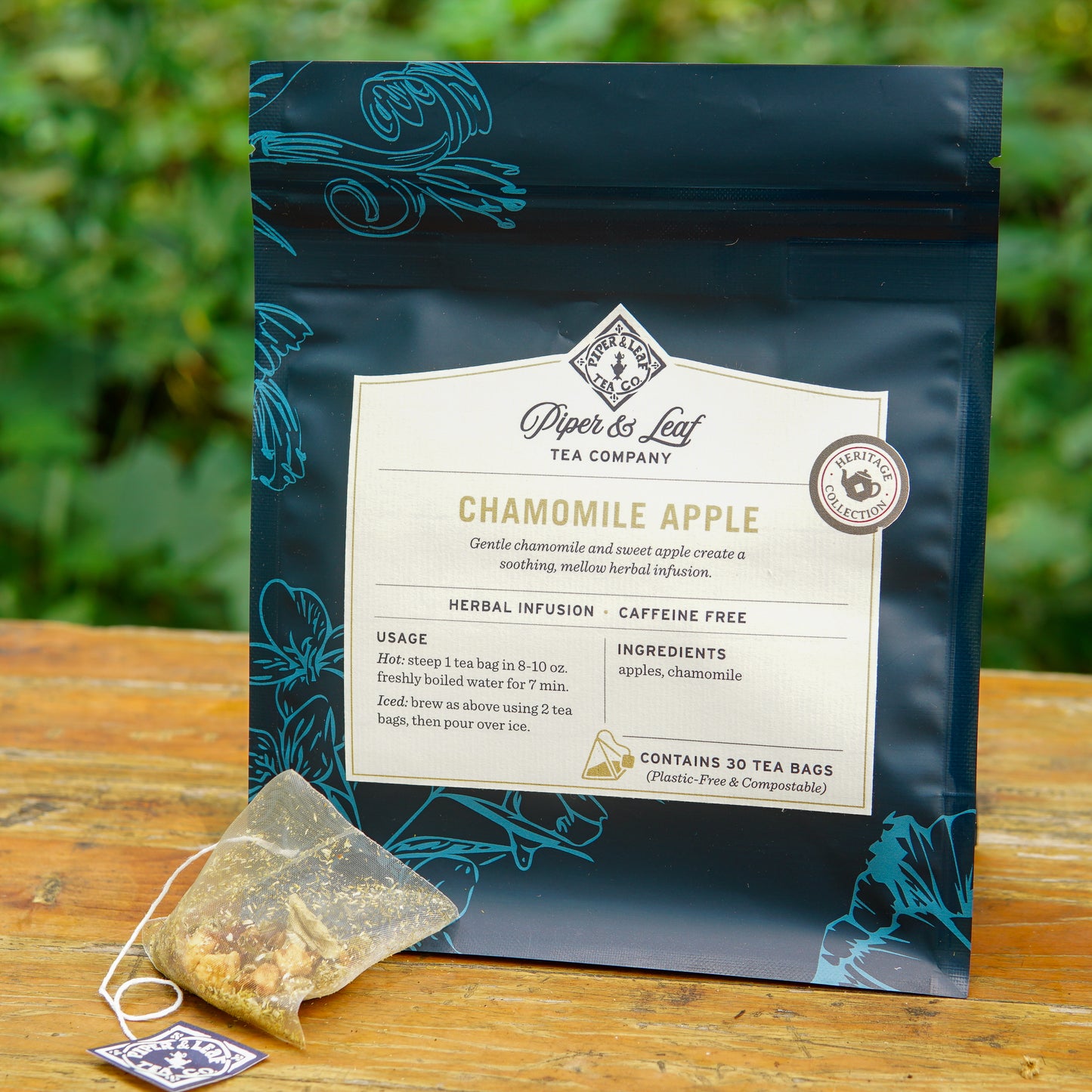 A package of Piper and Leaf Tea Co. Chamomile Apple 30ct Tea Bags rests outdoors; the packaging lists ingredients, usage instructions, and notes that it contains 30 plastic-free tea bags of this caffeine-free herbal infusion.