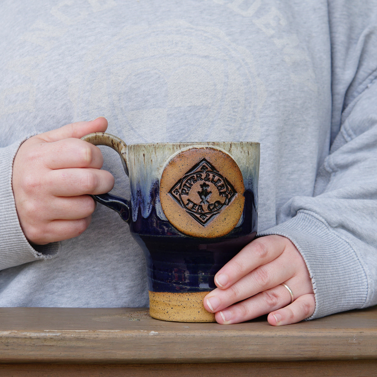 Person in a light gray sweatshirt holds a large Pottery Travel Mug featuring the Piper and Leaf Tea Co. logo, resting on a wooden surface.