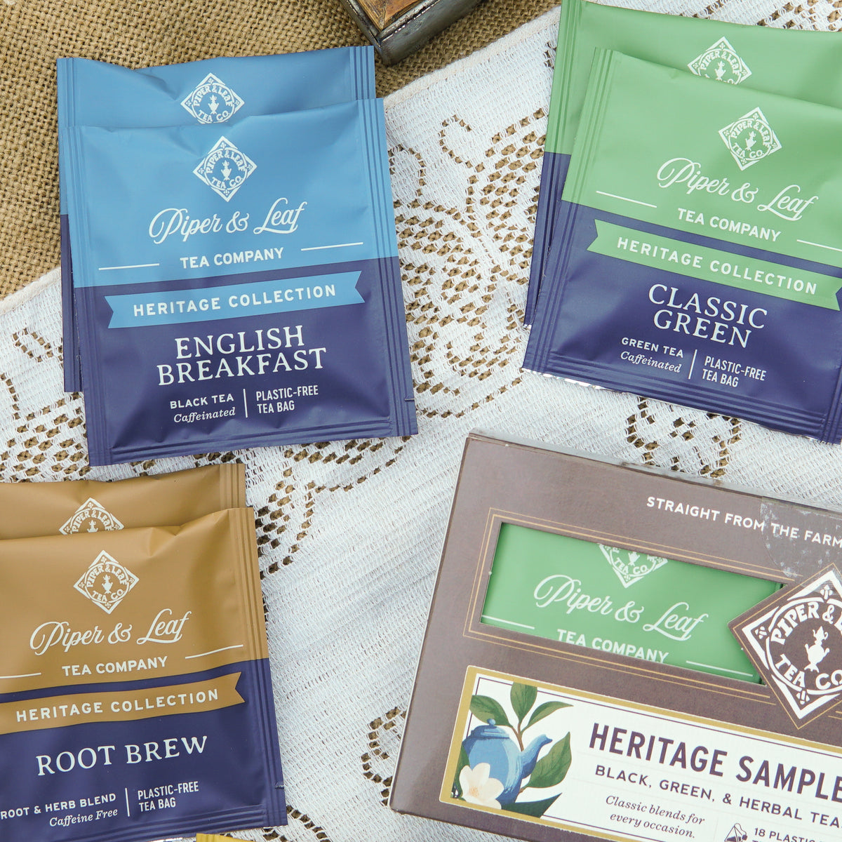 A selection of Piper and Leaf Tea Co. Heritage Collection premium tea blends—such as English Breakfast, Classic Green, and Root Brew—is arranged on a lace cloth alongside a Heritage Sampler box of 18 tea bags.