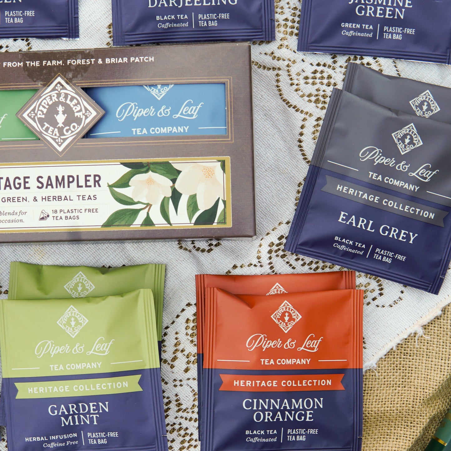 The Piper and Leaf Tea Co. Heritage Sampler - Box of 18 Tea Bags, featuring an assortment of premium blends, is elegantly displayed on a lace-covered surface.