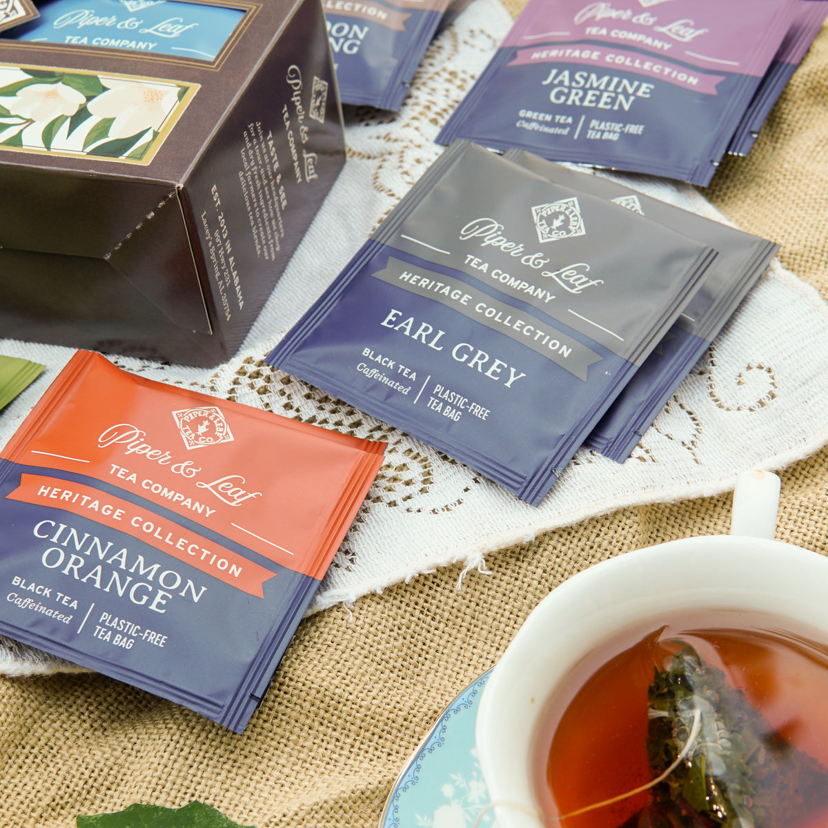 A Piper and Leaf Tea Co. Heritage Sampler—an 18-count box of premium tea bags—rests on the table alongside a cup of steeped tea, with several blends from the Heritage Collection, such as Earl Grey and Cinnamon Orange, displayed nearby.