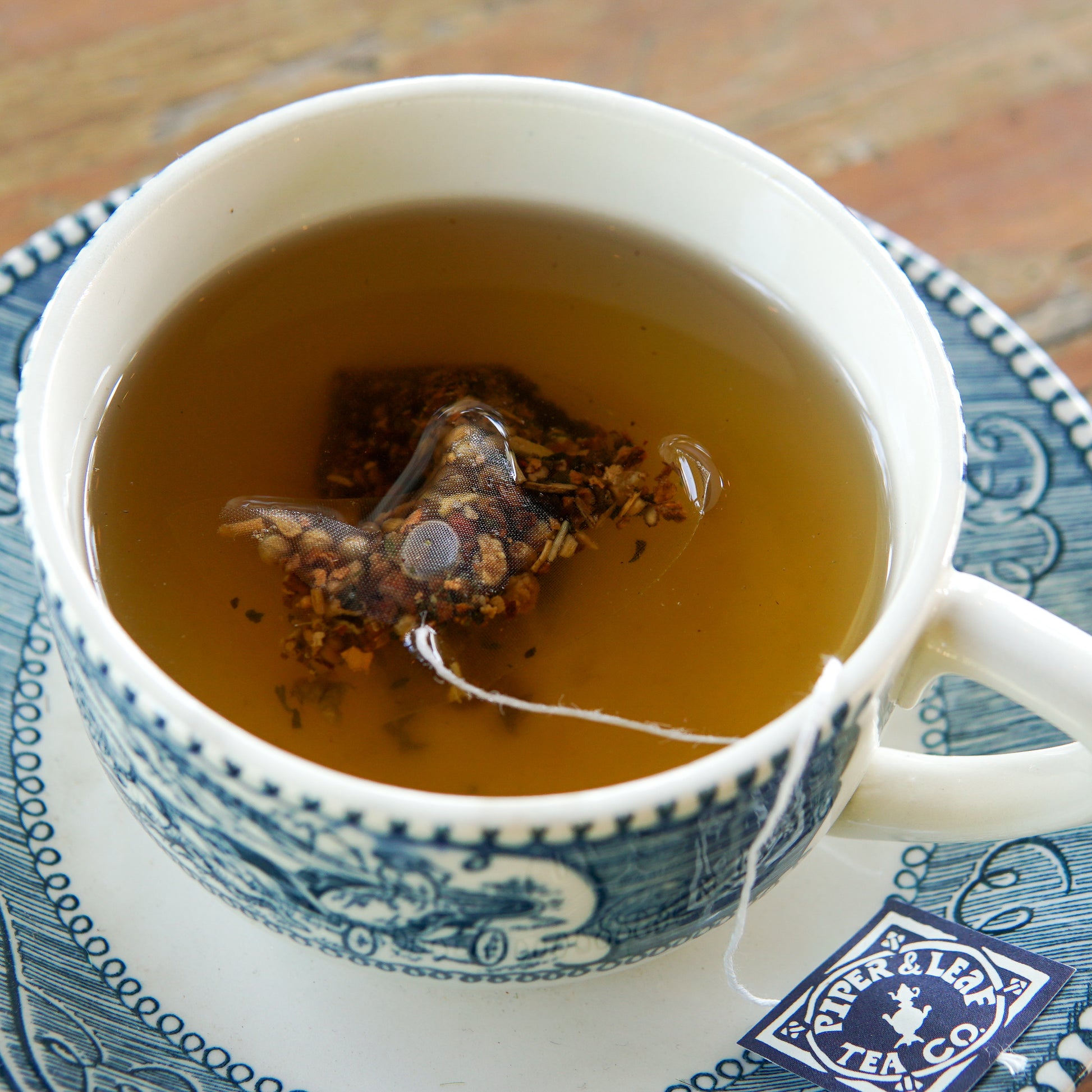 A teacup filled with freshly brewed tea—complete with a tea bag still steeping—rests on a matching saucer, displaying the Piper and Leaf Tea Co. logo from their Heritage Collection. The enticing aroma suggests this could be their unique Root Brew blend, available in a box of 50 individually wrapped tea bags.
