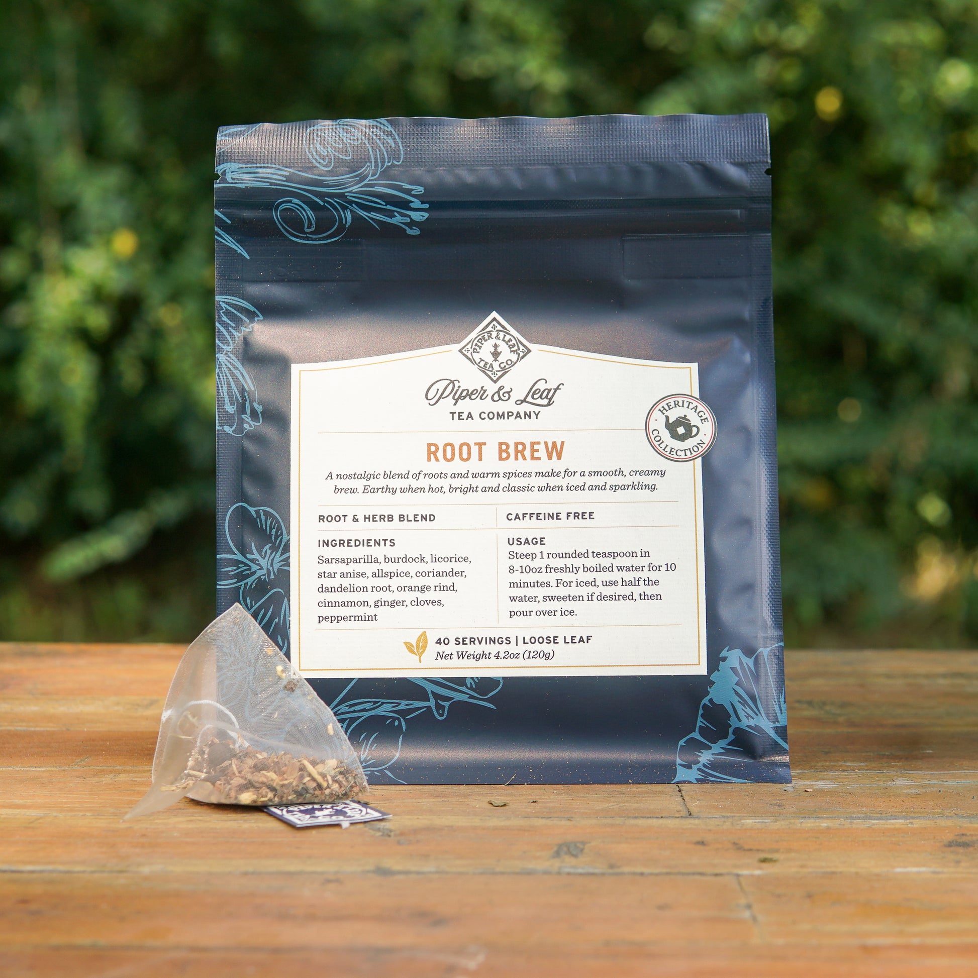 A package of Piper and Leaf Tea Co. Root Brew from the Heritage Collection sits on a wooden surface outdoors, with a caffeine-free herbal tea pyramid sachet in front of it and greenery blurred in the background.