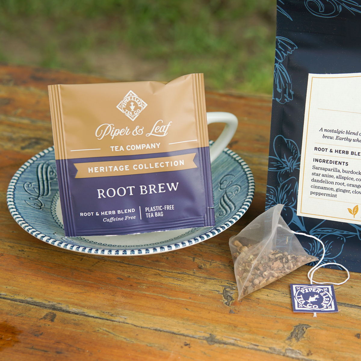 A Piper and Leaf Tea Co. Root Brew 50ct Individually Wrapped Tea Bag and its packaging rest on a blue plate beside a cup. A single sachet of this caffeine-free herbal tea, along with part of its package, can also be seen on the wooden surface, evoking classic root beer tea flavors.