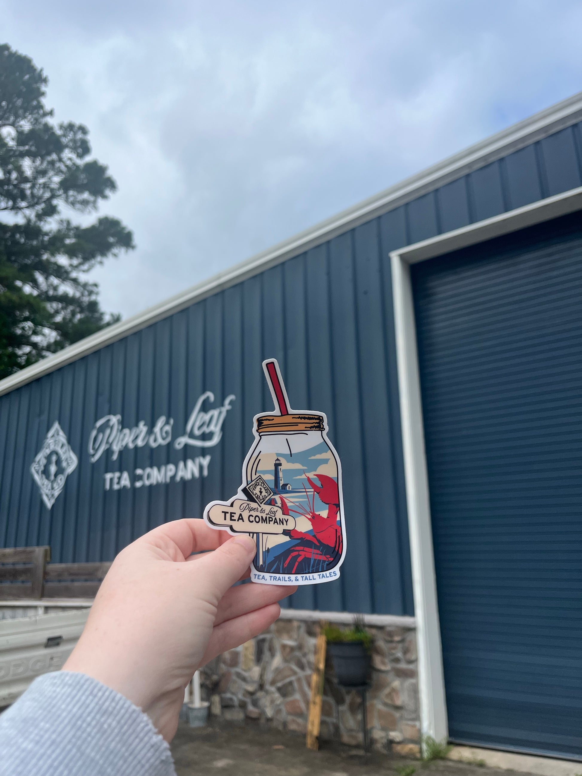 A hand holds a Nor'Easter Adventure Sticker from Piper and Leaf Tea Co. in front of the company’s blue metal building with a closed garage door.