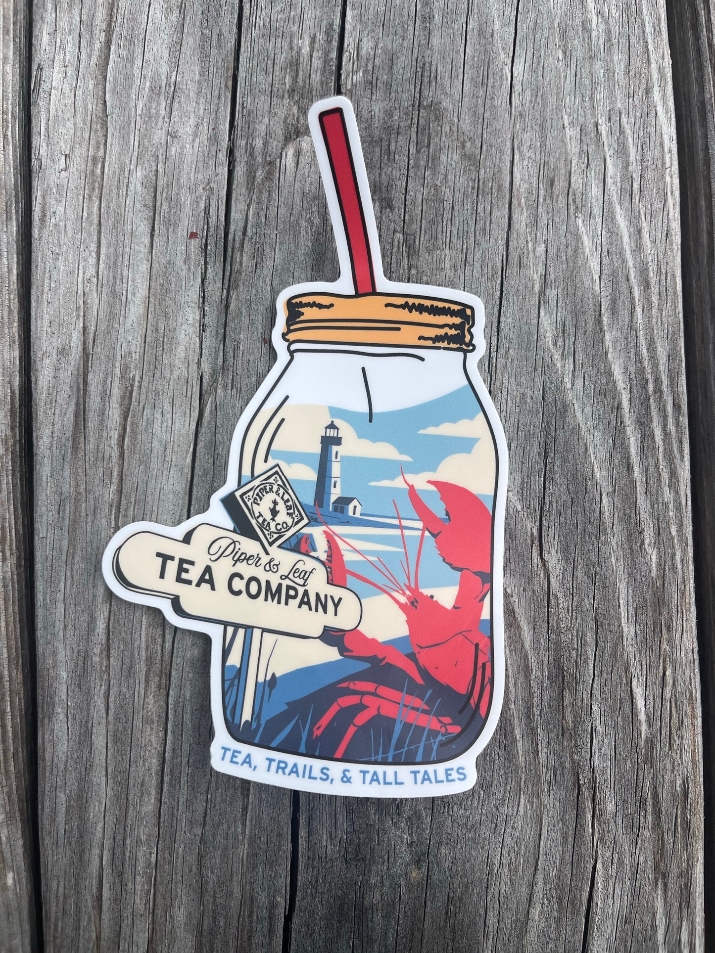 The Nor'Easter Adventure Sticker by Piper and Leaf Tea Co. features a mason jar with tea, a lighthouse, and a red lobster. “Piper’s Ley Tea Company” and “Tea, Trails, & Tall Tales” are printed on a weathered wood background.