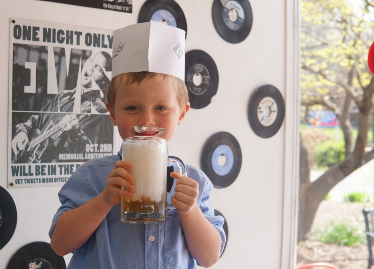 A young boy in a paper hat sips Piper and Leaf Tea Co.’s Root Brew, foam on his lip, standing by a wall lined with vinyl records and a concert poster.