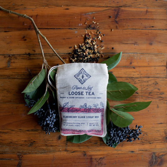 The Piper & Leaf Tea Co. Elderberry Elixir Muslin Bag of Loose Tea (15 servings) sits on a wooden surface with green leaves, berries, and dried flowers—ideal for making an immune-boosting tonic.