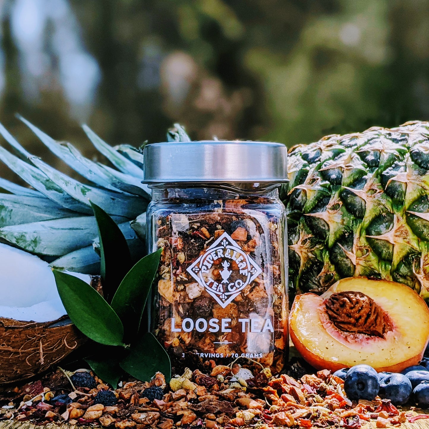 A Sweet Dixie Glass Jar of Loose Leaf Tea from Piper & Leaf Tea Co. sits among tropical fruits like pineapple, coconut, and peaches, surrounded by green leaves and blueberries.